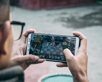A person is holding a smartphone horizontally, immersed in playing a complex strategy game. The focus is on the game screen, which displays an intricate board setup with various characters and elements.